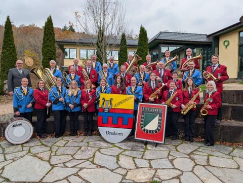 Ein Platz mit einem Orchester, das unter einem überdachten Bereich auftritt. Im Vordergrund steht ein Banner mit dem Logo von Ettelscheid.