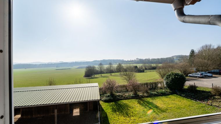 Un paysage vaste avec des champs verts et des collines douces. Au premier plan, on peut voir une structure avec un toit en métal et des arbres.