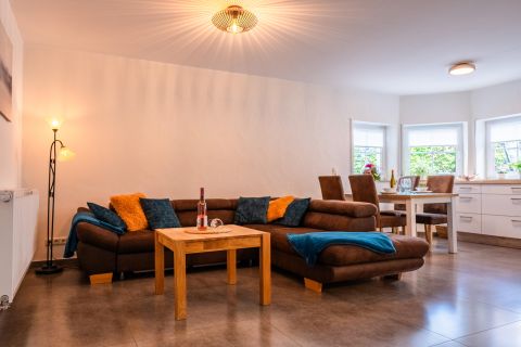 A modern living room with a brown sofa and colorful cushions. In the background, there is a dining area with a table and chairs.