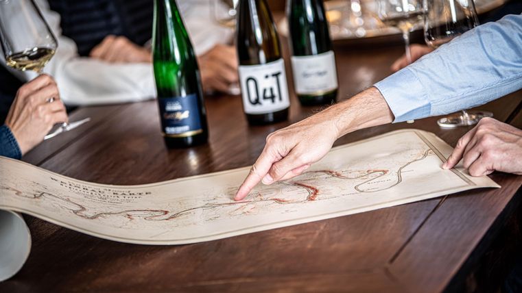 Un groupe de personnes regarde une grande carte et pointe un certain endroit. En arrière-plan, il y a quelques bouteilles de vin et des verres sur une table.