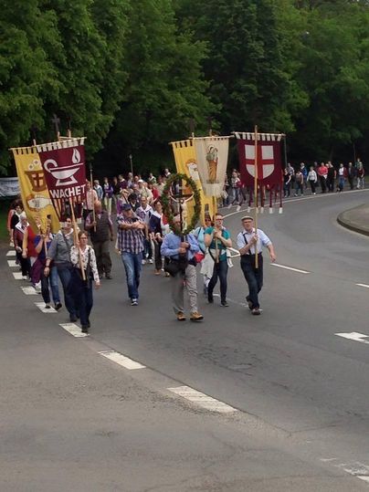 Eine Gruppe von Menschen marschiert auf einer Straße, während sie Banner tragen. Bäume umgeben die Szene, und es scheint eine festliche Veranstaltung zu sein.