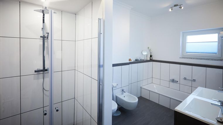 Ein modernes Badezimmer mit einer Duschkabine auf der linken Seite und einer Badewanne auf der rechten Seite. Die Wände sind hell gefliest und das Raumdesign ist schlicht und eleganter.