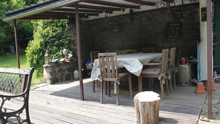 Een gezellige overdekte eetruimte in de buitenlucht met een tafel en stoelen. Omringd door groene planten en een houtstam als kruk.