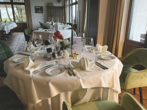 In de voorgrond een gedekte tafel met gevouwen servetten, wijnglazen en bestek ; In de achtergrond zie je de rest van de ruimte met tafels