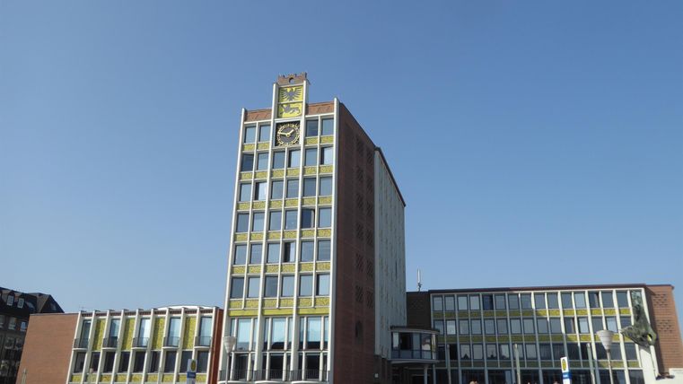 A modern building with a striking facade and a tall tower. People stand in front of it enjoying the sunny day.