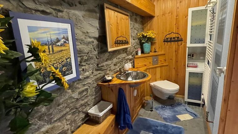 A cozy bathroom with wooden furniture and stone walls.
There is a sink with a mirror and decorations made of flowers.  