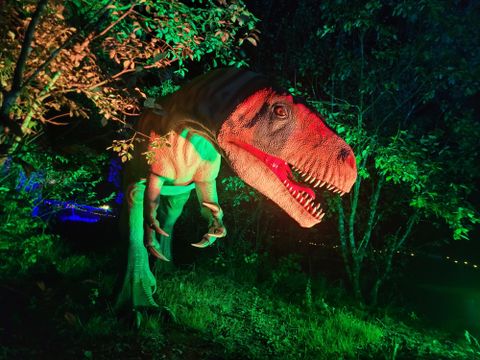 Un dinosaure réaliste, se tenant entre des arbres, illuminé par une lumière verte et rouge. La scène dégage une atmosphère mystique et palpitante.