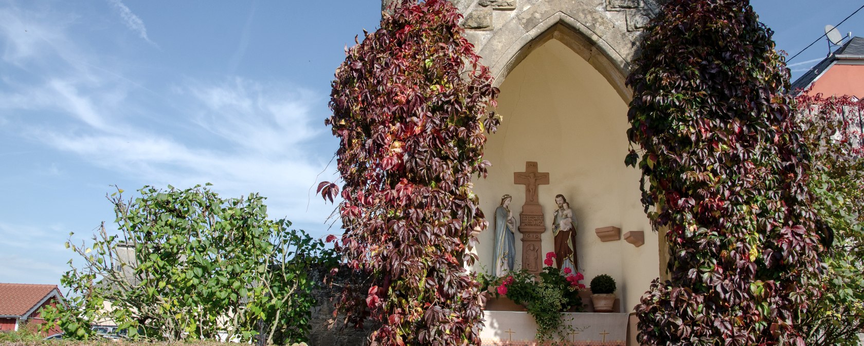 Une petite chapelle à Wolsfeld, entourée de plantes grimpantes rouges. A l'intérieur, on peut voir des statues religieuses. Le ciel est clair et bleu., © Tourist-Information Bitburger Land