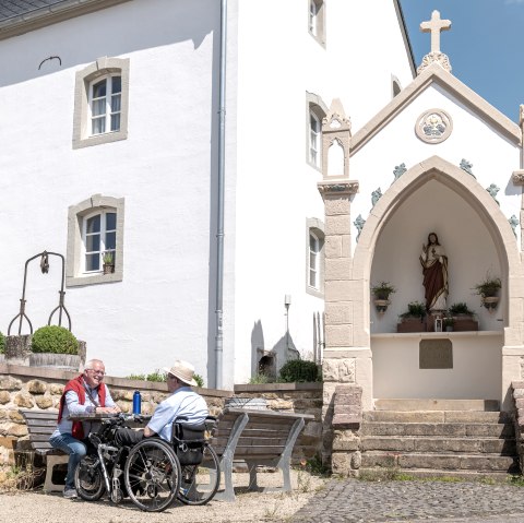 Zwei Männer, einer im Rollstuhl, sitzen auf einer Bank vor einem weißen Gebäude mit einer religiösen Statue in einer Nische. Sonniger Tag., © Naturpark Südeifel, Thomas Urbany