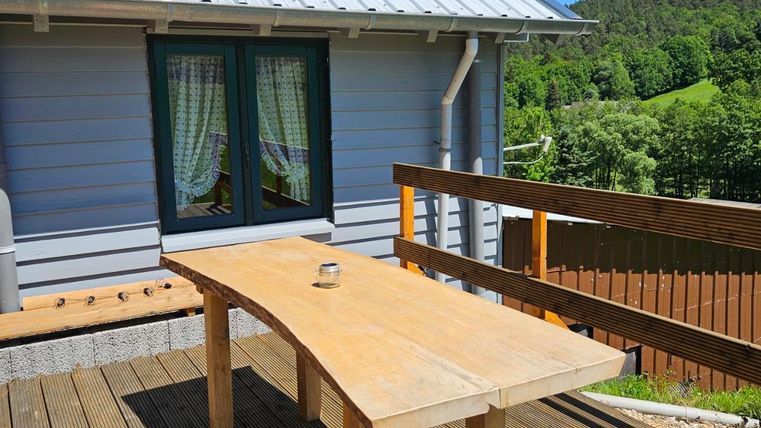 A wooden table is standing on a terrace in front of a gray house. The surroundings are green and sunny, with a clear blue sky.