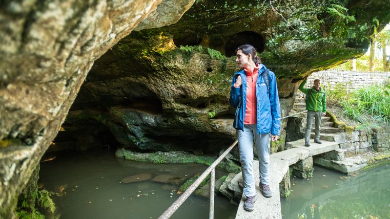 Twee mensen staan op een smalle loopbrug over een vijver, omgeven door rotsen en vegetatie.