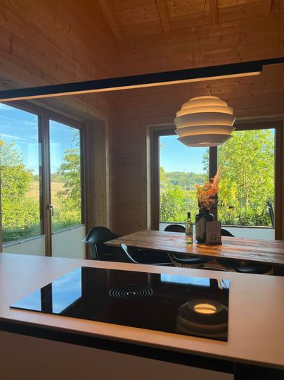 Eine moderne Küche mit einem Kochfeld und einem Essbereich. Große Fenster bieten einen Blick auf die grüne Landschaft.