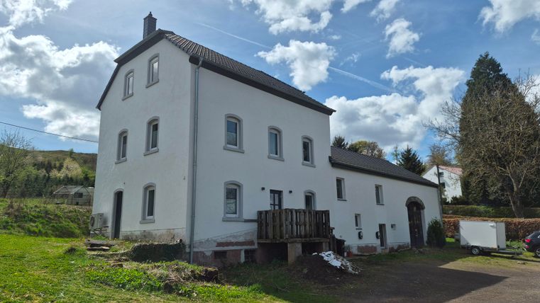 Een wit huis met een moderne gevel en een klein houten terras. De omgeving is groen met bomen en een helder blauwe lucht.