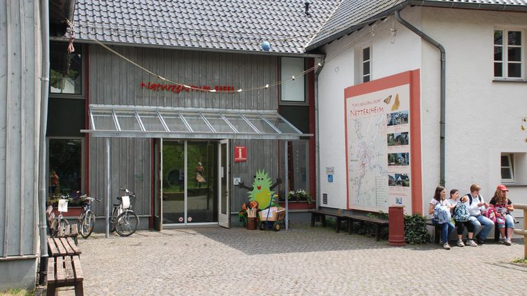 A modern building with an entrance area and a large window. Outside, several people are sitting on a bench.