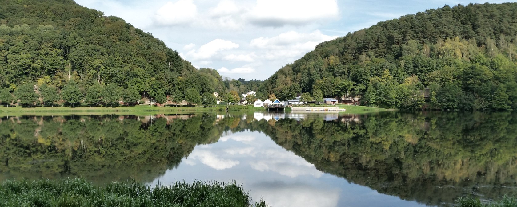 Panorama of the Bitburg reservoir with wooded hills, buildings on the shore and a clear reflection in the water under a blue sky., © TI Bitburger Land