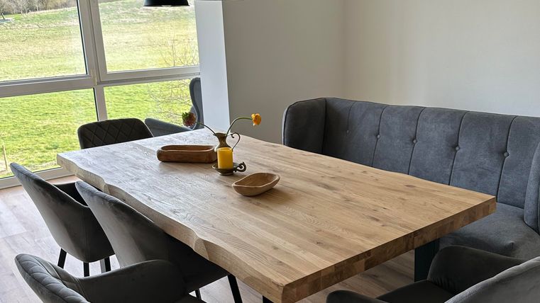 Ein modern eingerichteter Essbereich mit einem großen Holztisch und grau gepolsterten Stühlen. Durch die Fenster ist eine grüne Landschaft zu sehen.