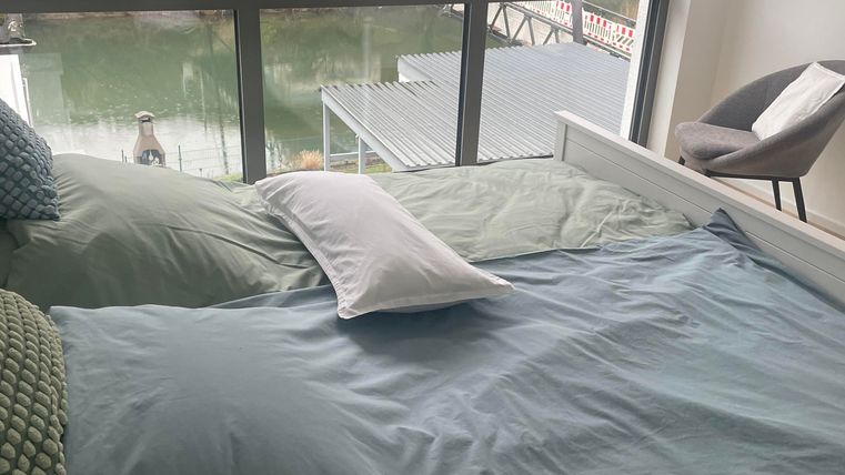 Ein modernes Schlafzimmer mit einem großen Fenster und Blick auf einen ruhigen Wasserkanal. Das Bett ist mit blauen Bettdecken und Kissen dekoriert.