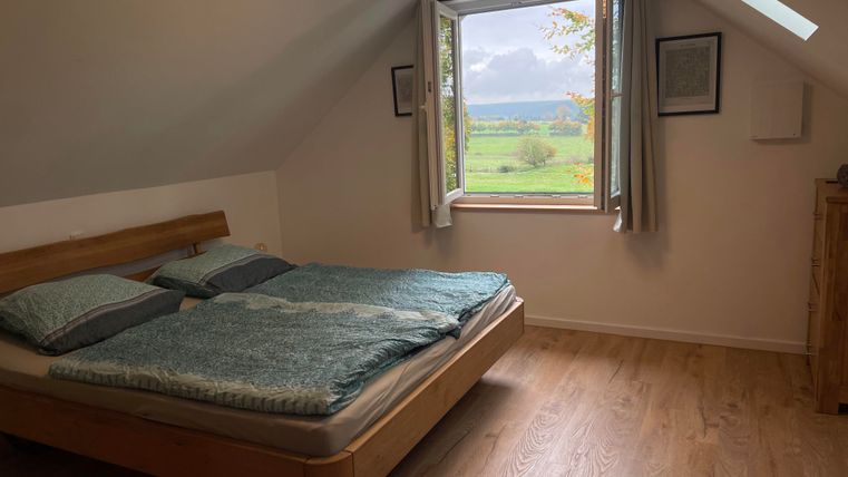 Ein gemütliches Schlafzimmer mit einem Doppelbett und sanften Farben. Das Fenster bietet einen Blick auf eine grüne Landschaft unter einem bewölkten Himmel.