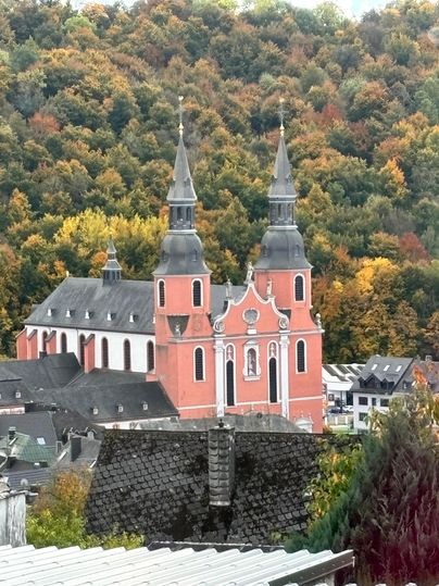 A church with two pointed towers stands in front of a colorful forest. The colors of the trees display autumn.