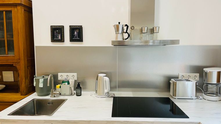 Une cuisine moderne avec des armoires blanches et un dosseret en acier inoxydable. Sur le plan de travail, divers appareils de cuisine tels qu'une bouilloire et une plaque de cuisson sont visibles.