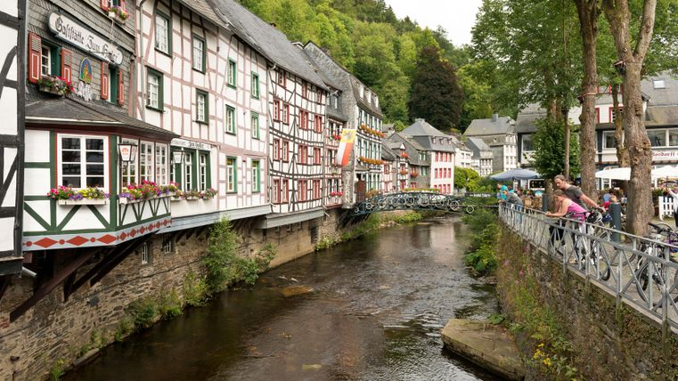 Vakwerkhuizen in Monschau aan een rivier met een brug en wandelaars.
