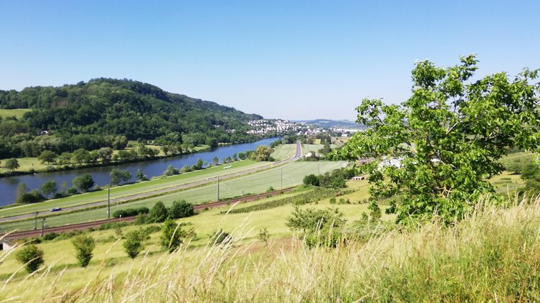 Een pittoresk landschap met een rivier, omgeven door glooiende heuvels en groene weiden. De lucht is helder en blauw, wat de idylle versterkt.