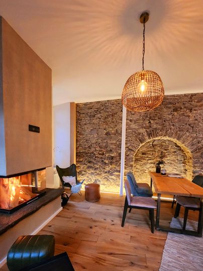 A cozy living area with an open fireplace and a large wooden table. The walls are clad in natural stone, and a beautiful lamp provides atmospheric lighting.