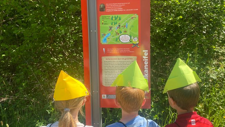 Drei Kinder mit bunten Hüten und grünen Rucksäcken stehen vor einem Schild des Märchenpfads Jünkerath.