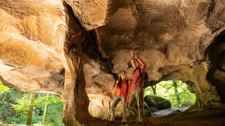 Deux randonneurs se tiennent dans une grotte rocheuse au plafond voûté et regardent vers le haut.
