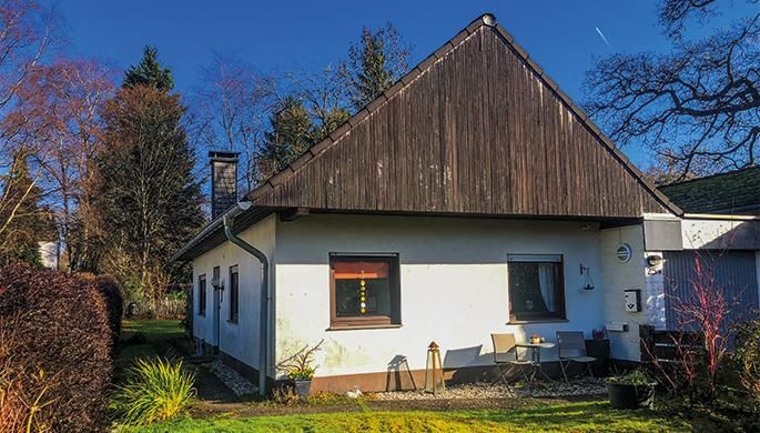 Ein einfaches Haus mit einem geneigten Dach und einem kleinen Garten. Es ist von Bäumen umgeben und hat einen klaren blauen Himmel im Hintergrund.