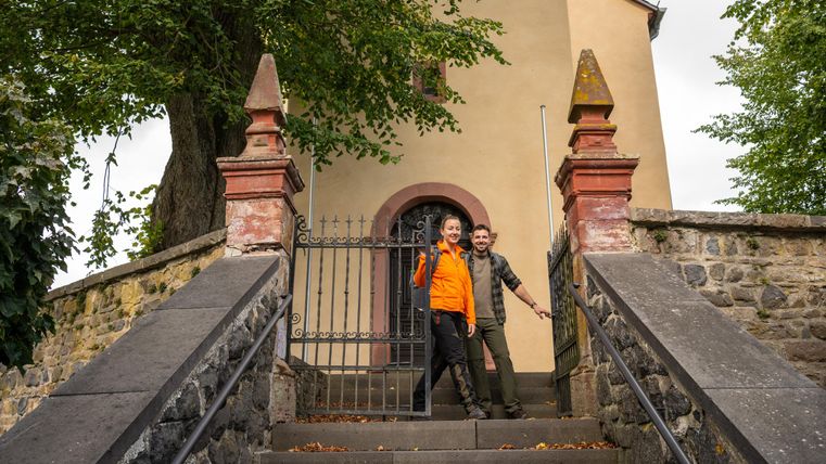 Two people are standing on a staircase in front of a building. The surroundings are green and inviting, perfect for outdoor activities.