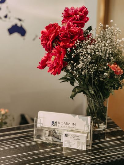 Un joli bouquet de fleurs avec des pivoines rouges et de délicates fleurs blanches est posé sur une table. En dessous, des cartes de visite de l'entreprise KOMM'IN sont visibles.