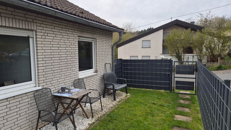 Een gezellige buitenruimte met een tafel en stoelen voor een huis. Het groene gras en de omheining zorgen voor een rustige sfeer.