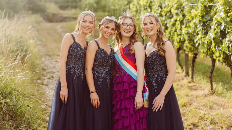 Four young women in elegant evening gowns pose smiling in a vineyard. The atmosphere is festive and cheerful.