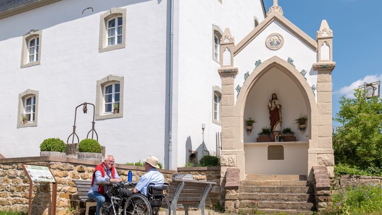 Twee mannen zitten op een bankje voor een wit gebouw met een religieus beeld in een nis.