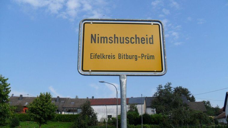 Ein Ortsschild von Nimshuscheid im Eifelkreis Bitburg-Prüm. Die Umgebung ist ländlich mit grünen Wiesen und Häusern im Hintergrund.
