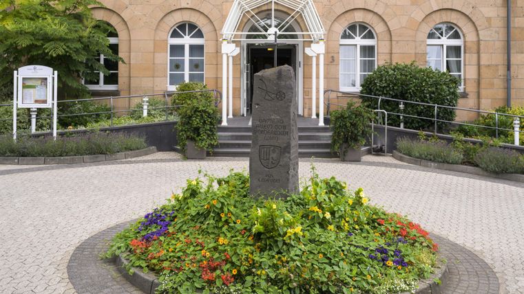 Een mooi hotel met een uitnodigende gevel en een bloeiend bloemenbed vooraan. De ingang is omgeven door bomen, wat een vredige sfeer creëert.