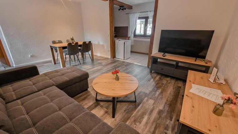 Ein gemütliches Wohnzimmer mit einem grauen Sofa und einem runden Holztisch. Im Hintergrund steht ein Fernseher und ein Esstisch.