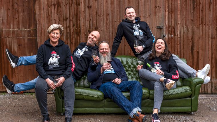 Eine Gruppe von fünf Personen sitzt und lehnt sich entspannt auf einer grünen Couch. Sie lachen und halten Getränke in der Hand, während sie lässige Hoodies tragen.