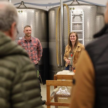 Twee personen staan in een instelling met grote zilveren tanks. Ze glimlachen en lijken een presentatie of gesprek te voeren.