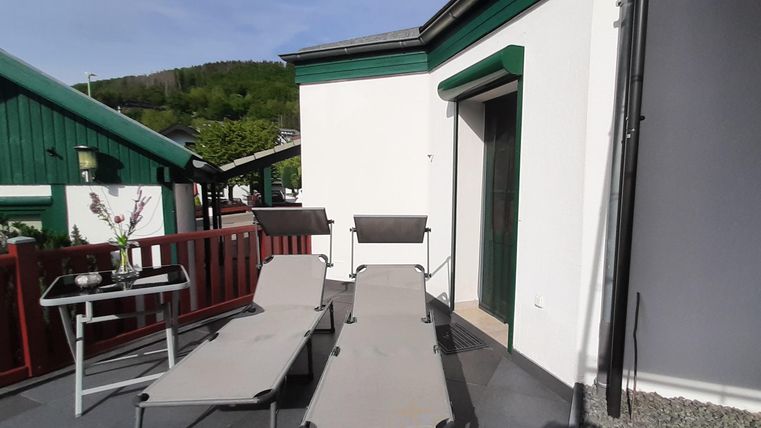 Une terrasse ensoleillée avec deux chaises longues et une petite table. En arrière-plan, on peut voir des bâtiments verts et un paysage agréable.