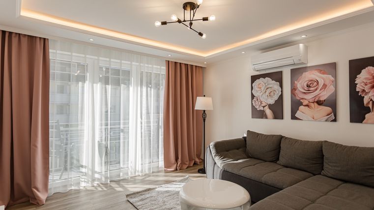 A modern living room with a gray sofa and a round table. Pictures of roses hang on the wall, and large windows let in a lot of light.