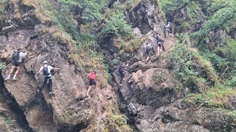 A group of people climbs along steep rocks. The surroundings are green and wooded, with a water source nearby.