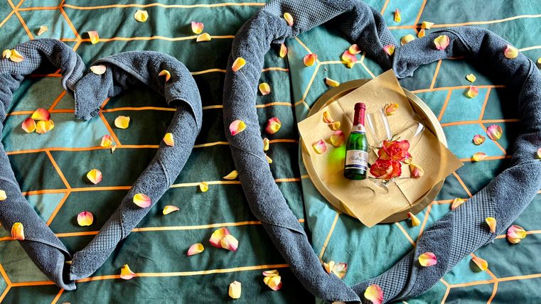 Two towel hearts lie on a bed, surrounded by rose petals. In the middle is a small plate with flowers and a bottle of champagne.