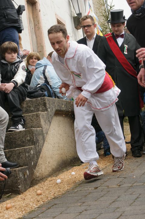 A man in white attire is walking down a staircase while balancing eggs. Around him, spectators are watching with anticipation.
