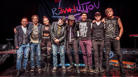 Een groep van zes muzikanten poseert in een gezellige ruimte. Ze staan en zitten rondom een rode fauteuil en dragen casual kleding.