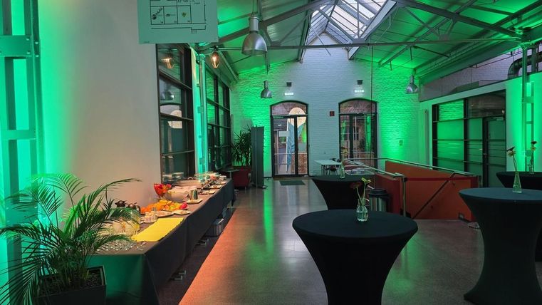 Een moderne evenementenruimte met groene verlichting. Op een tafel zijn verschillende snacks en dranken gepresenteerd.
