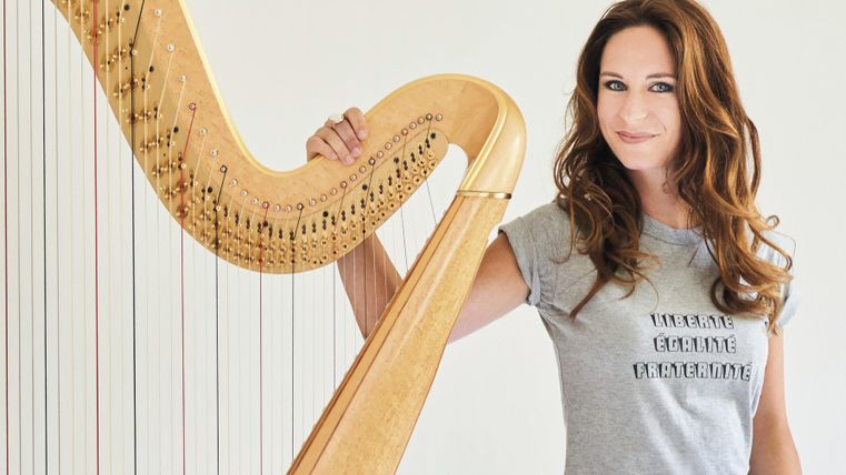 Eine Frau steht neben einer Harfe und lächelt. Sie trägt ein graues T-Shirt mit einem Schriftzug.