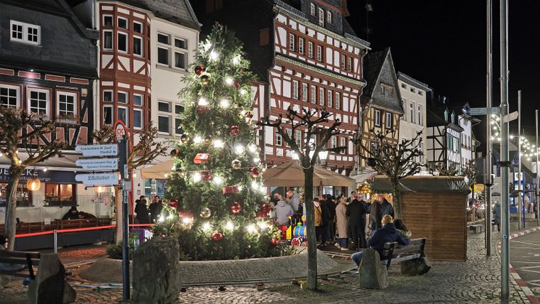 A festively illuminated street with a large Christmas tree. People gather in the twilight among the nicely decorated buildings.