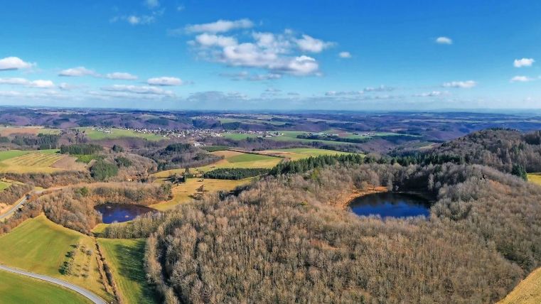 Een schilderachtig landschap met zachte heuvels en beboste gebieden. Op de voorgrond zijn twee ronde waterpartijen zichtbaar, omringd door weiden en bomen.